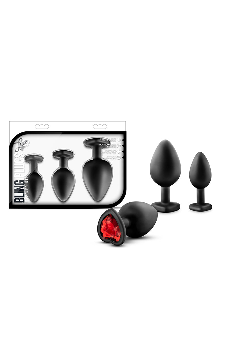 Three matte black silicone anal plugs with red faceted heart gem bases, beside the Bling Plugs Trainer Kit package.