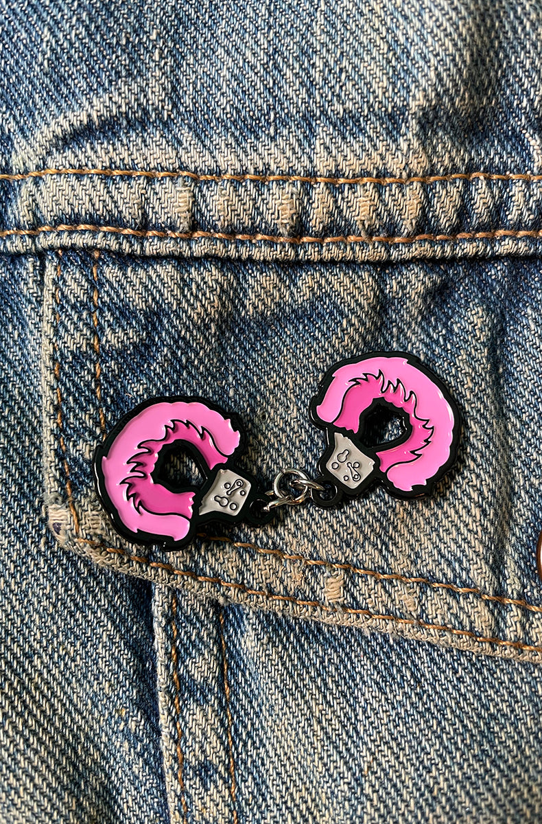Hot-pink fuzzy handcuffs enamel pin with two cuffs joined by a tiny silver chain, attached to a denim jacket pocket.