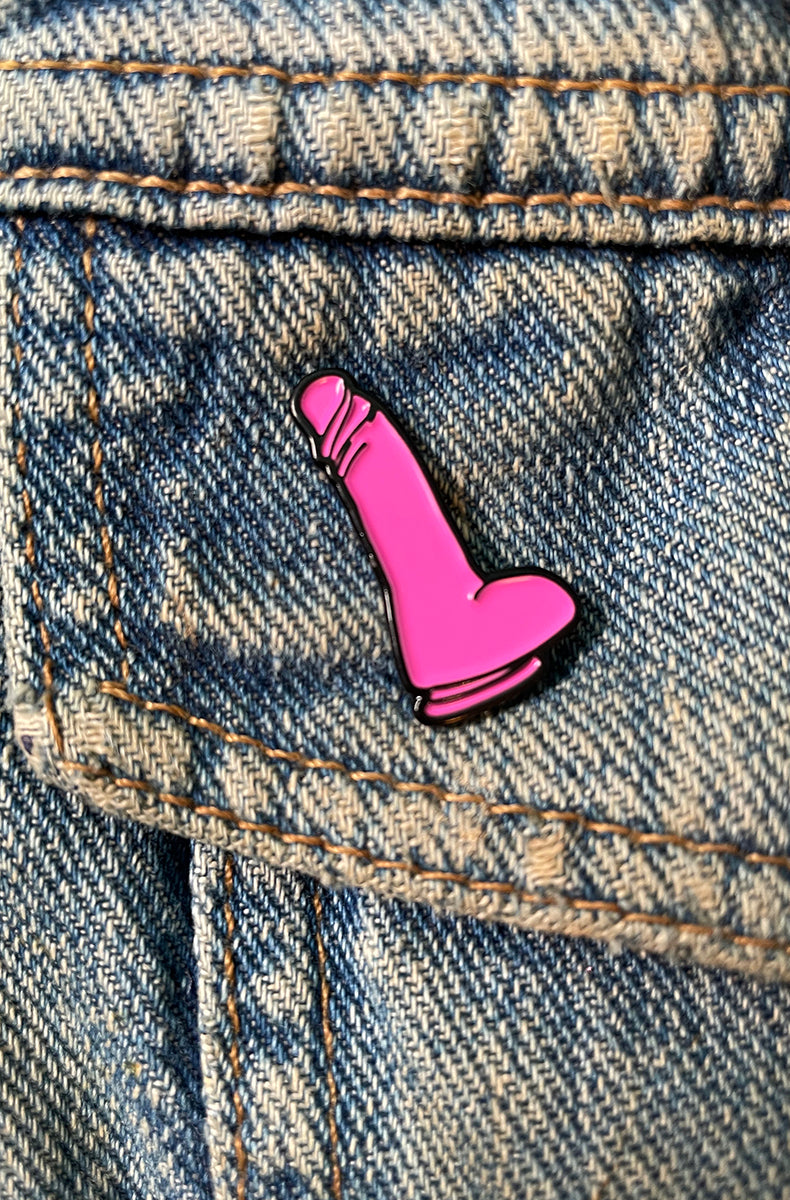 Hot-pink enamel pin shaped like a small dildo with darker pink shading lines, attached to a denim jacket pocket.