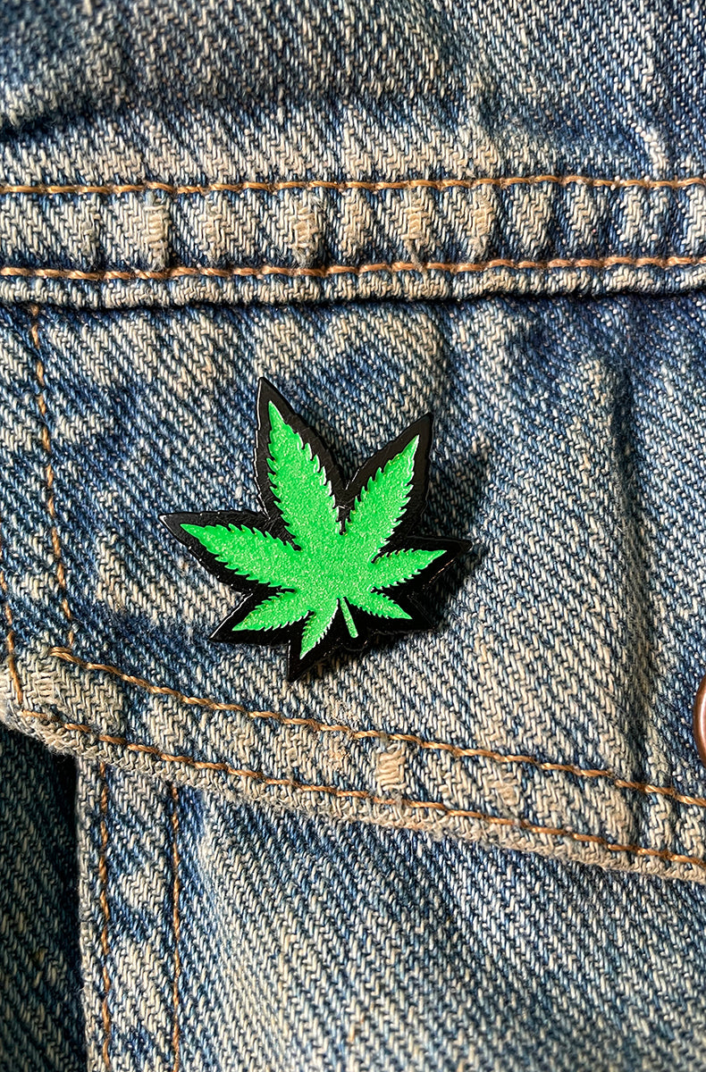 Bright green cannabis leaf enamel pin with glossy black outline, attached to a denim jacket pocket.