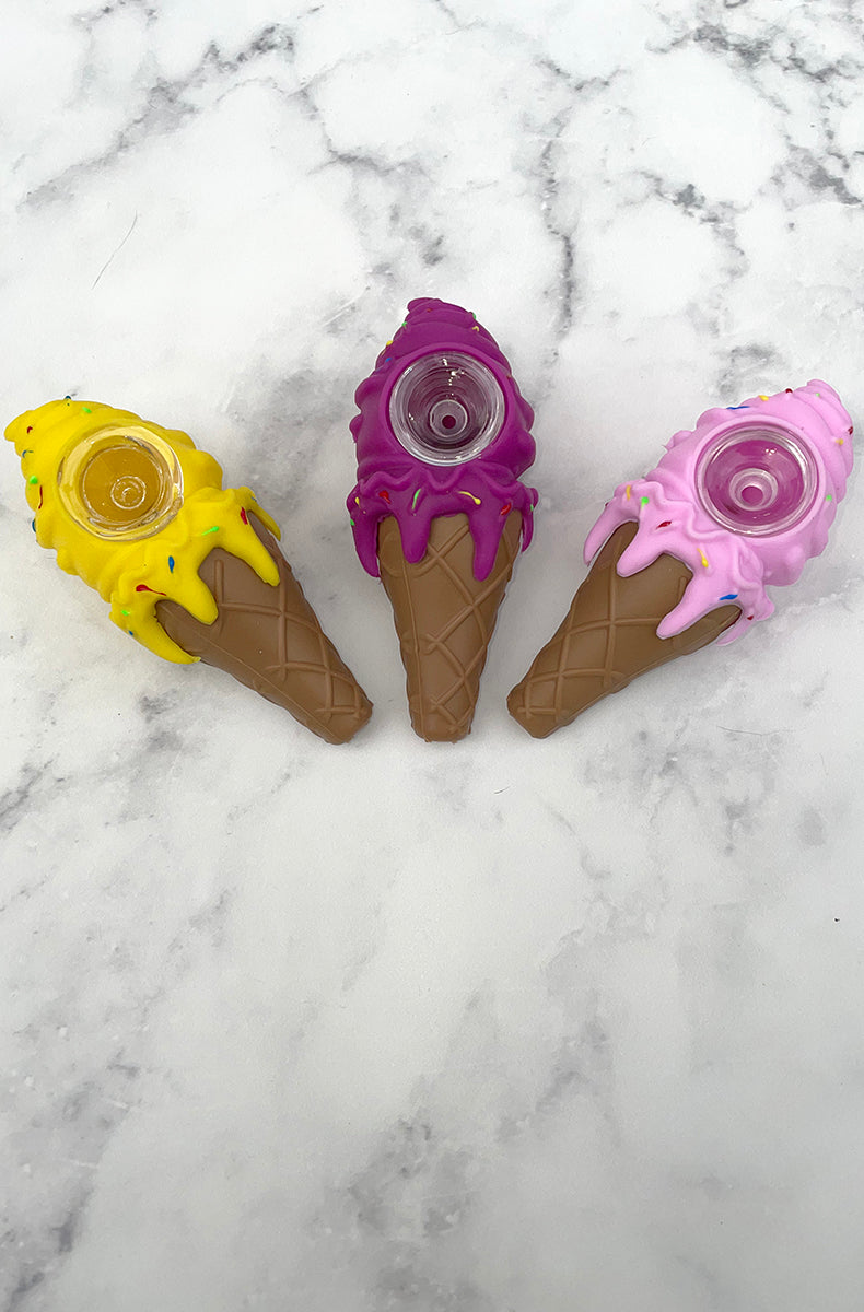 Three PIECEMAKER ice cream cone silicone pipes in yellow, purple, and pink with sprinkle accents and glass bowls, on marble.