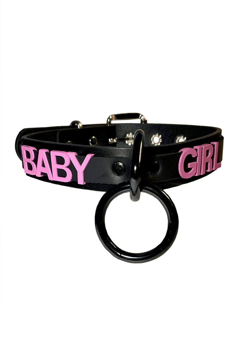 Black vegan-leather collar with "BABY" and "GIRL" in bold pink lettering and a large black metal O-ring at the front center.