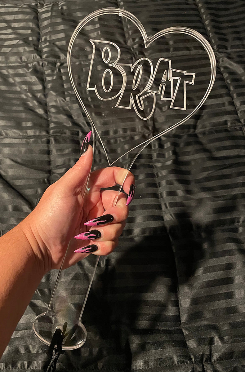Clear cast acrylic heart-shaped paddle with BRAT engraved in graffiti-style block letters inside the heart outline.