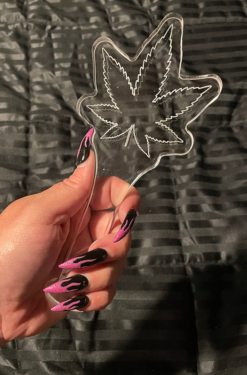 Clear cast acrylic paddle shaped like a cannabis leaf with an etched outline, held in a hand on a dark satin background.