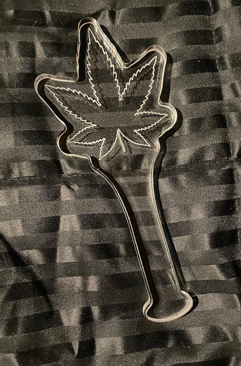 Front view of the clear acrylic cannabis-leaf paddle laid flat on a black striped satin background.