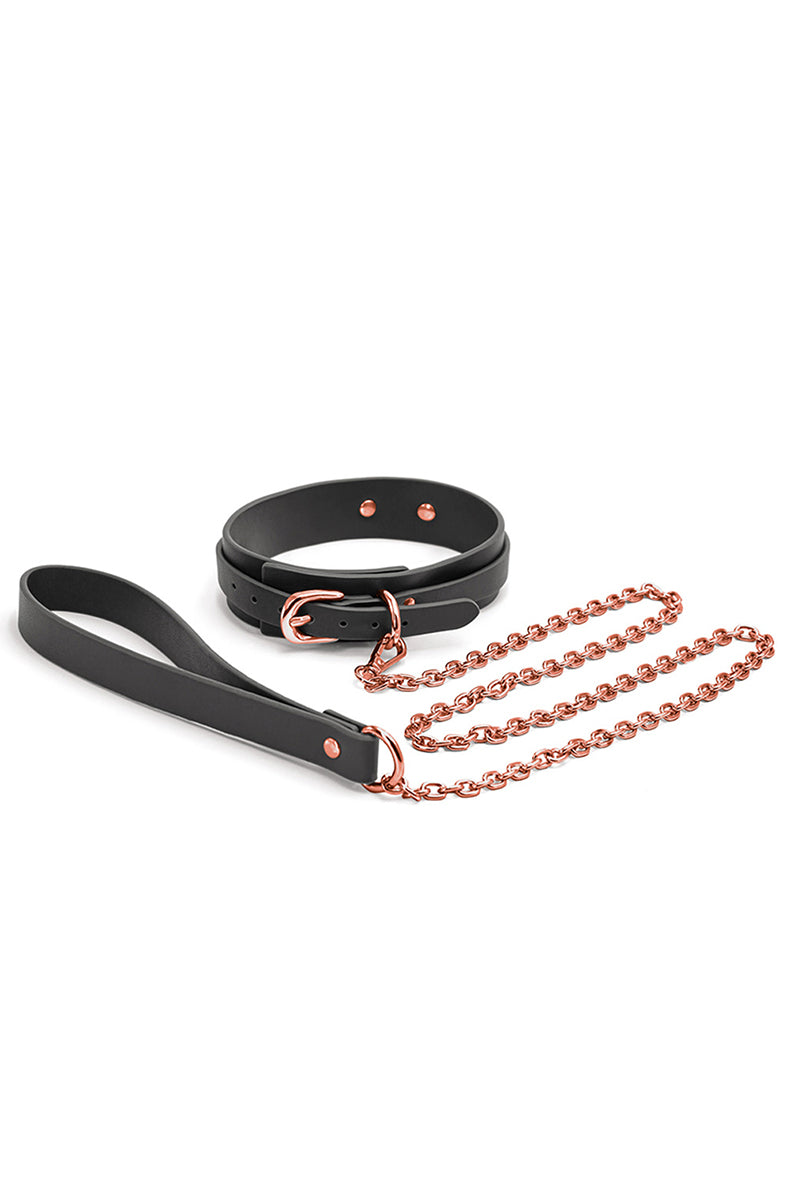 Narrow black synthetic-leather collar with rose-gold buckle and D-ring, with a rose-gold chain leash on a black hand loop.