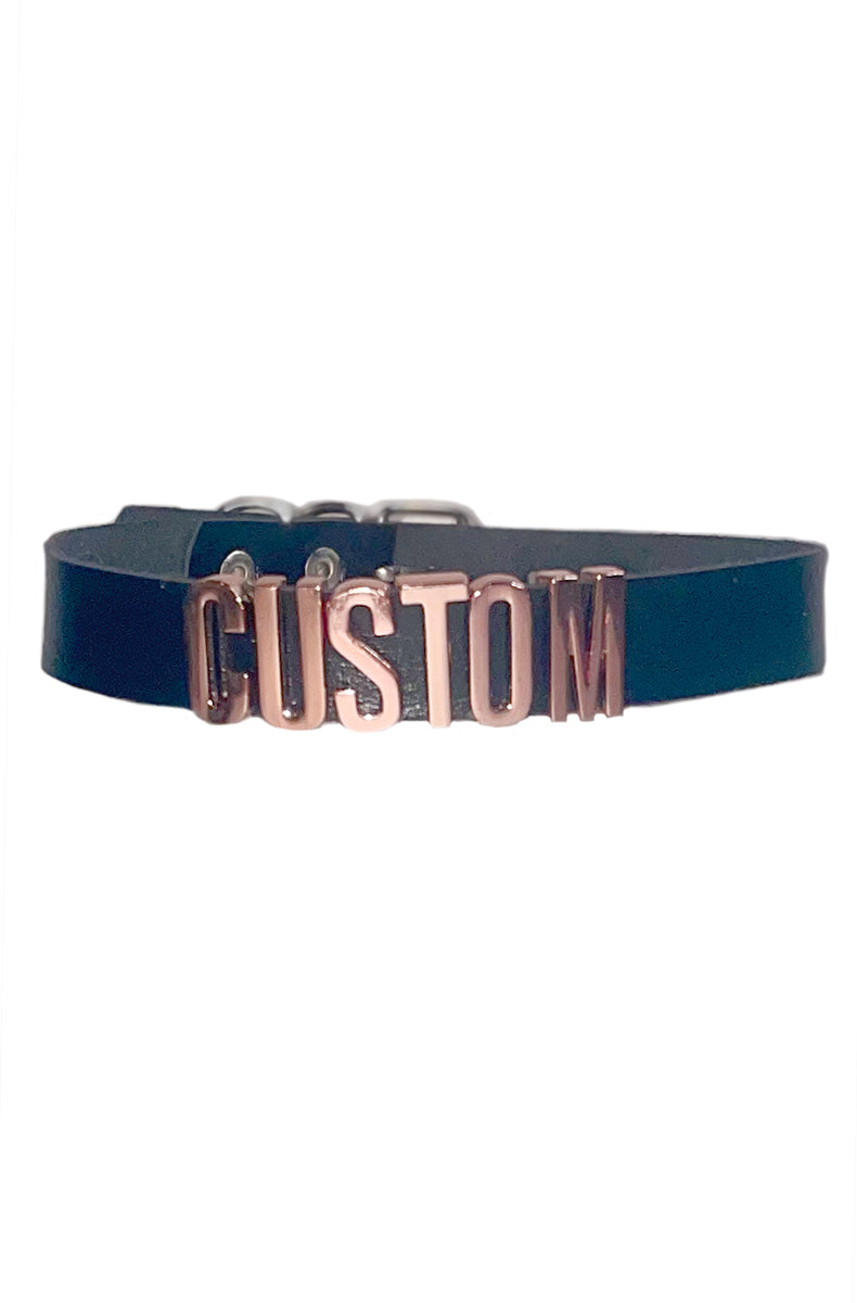 Black vegan leather collar with raised rose-gold metal letters spelling CUSTOM as a sample, with silver buckle and D-rings at the back.