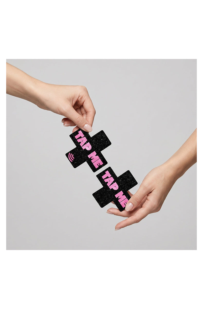 Two black glitter TAP ME cross pasties held between fingertips