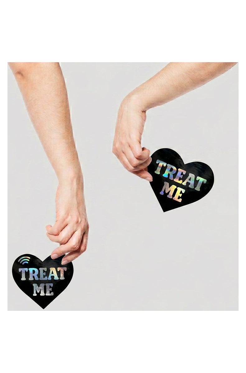 Two black TREAT ME heart pasties held between fingertips