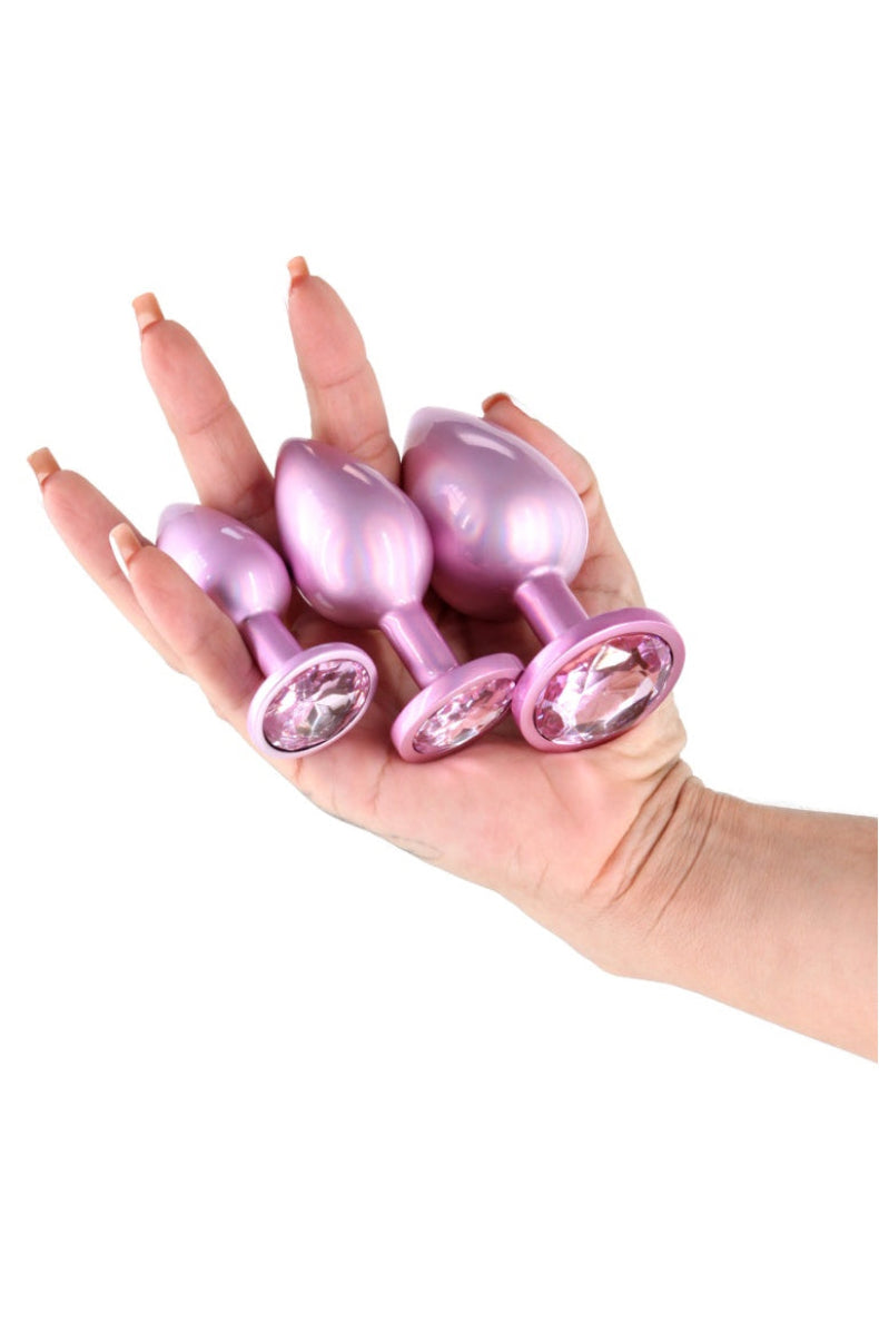 Three graduated pink metal butt plugs held in one hand for scale, each with a pale pink gem base