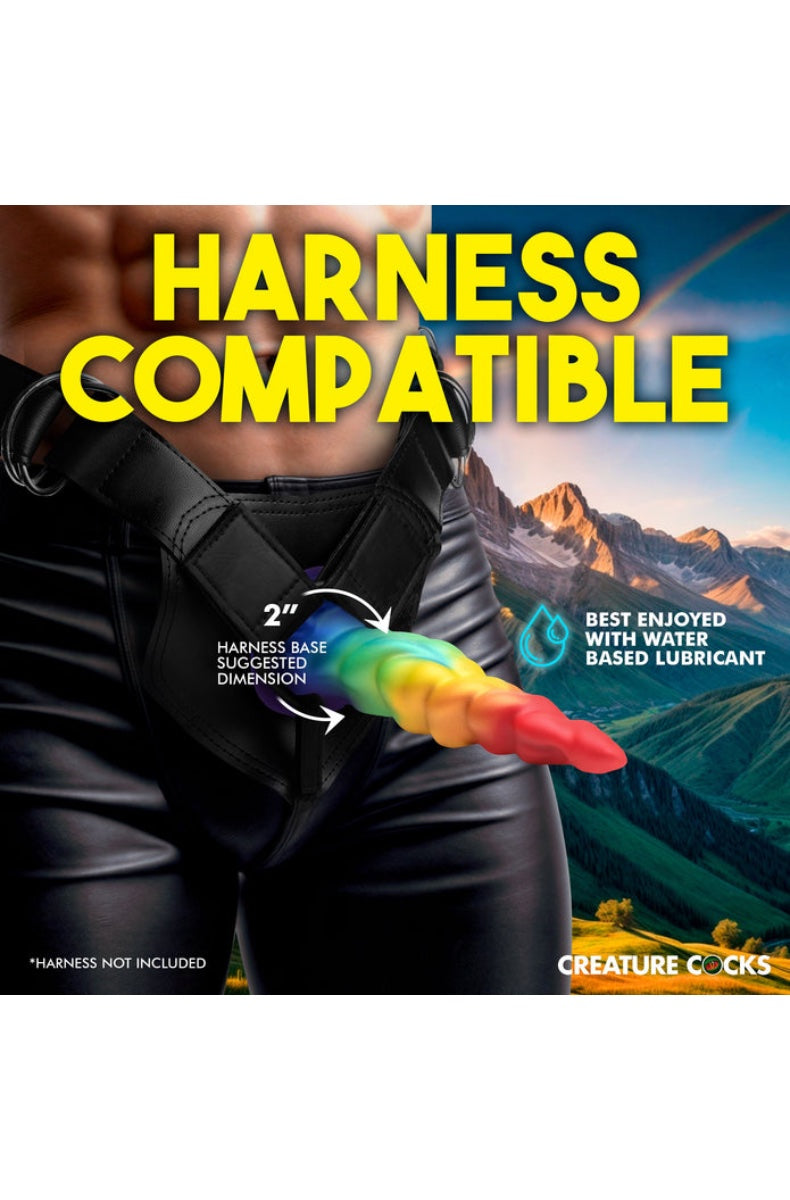 Model wearing black leather harness with rainbow dildo positioned to show 2 inch harness compatibility against mountain landscape