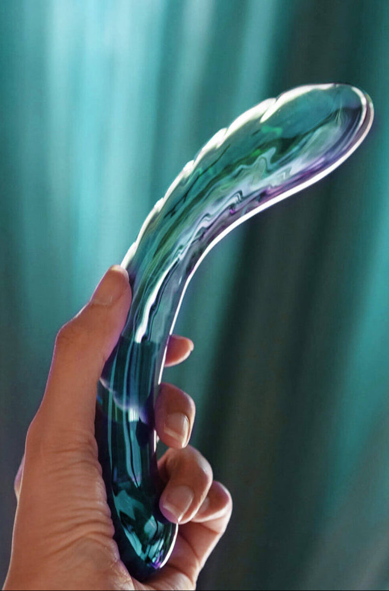 A hand holding the teal iridescent glass dildo in front of a teal curtain, showing the curved shaft and ribs.