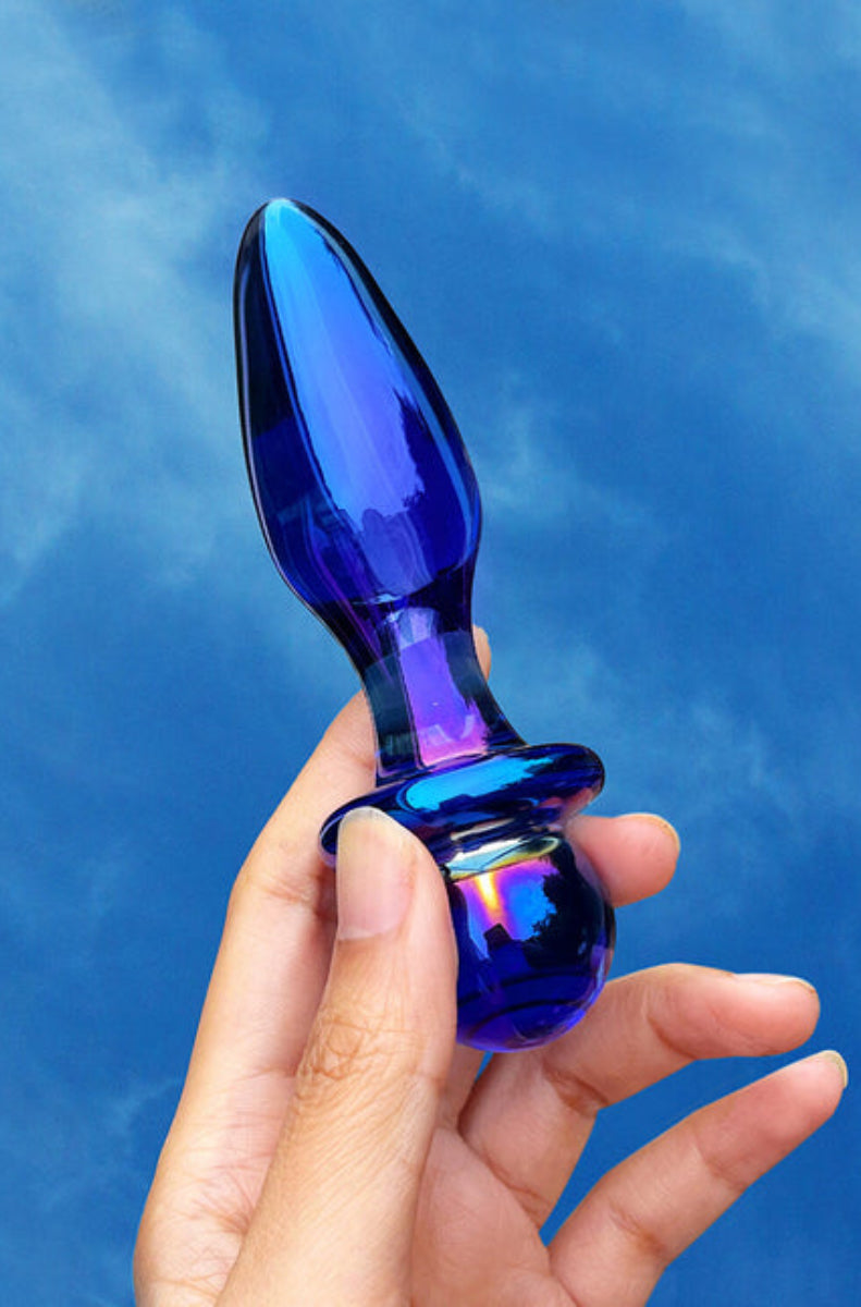 A hand holding the blue iridescent glass plug against a blue sky, showing the teardrop body and flared base.