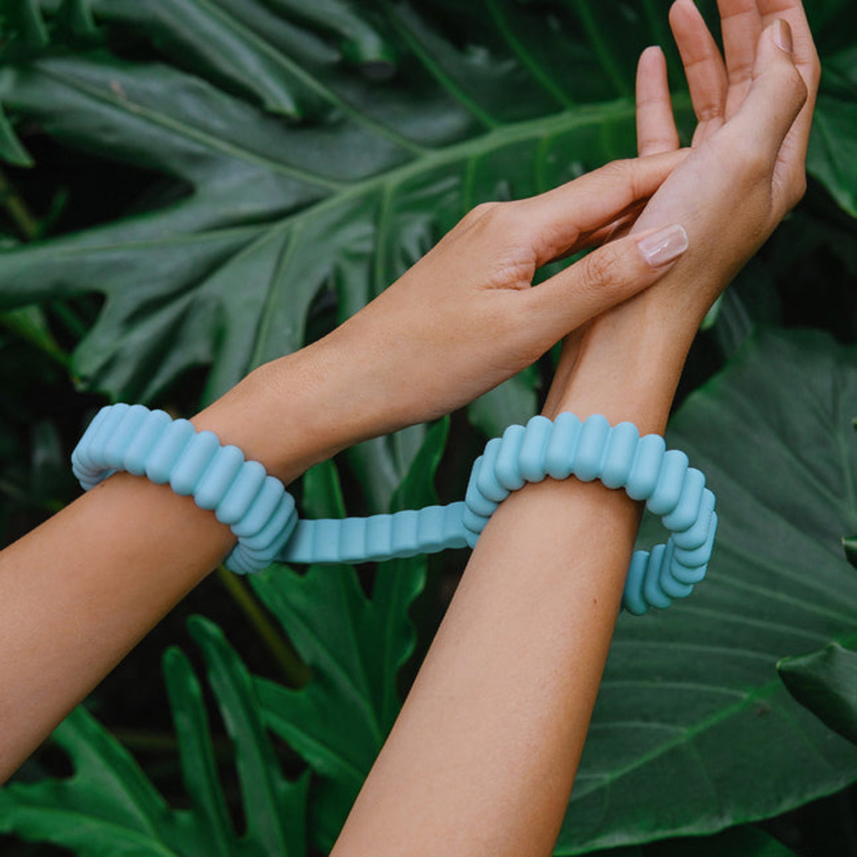 A model wearing the mint silicone cuffs on both wrists in front of tropical leaves.