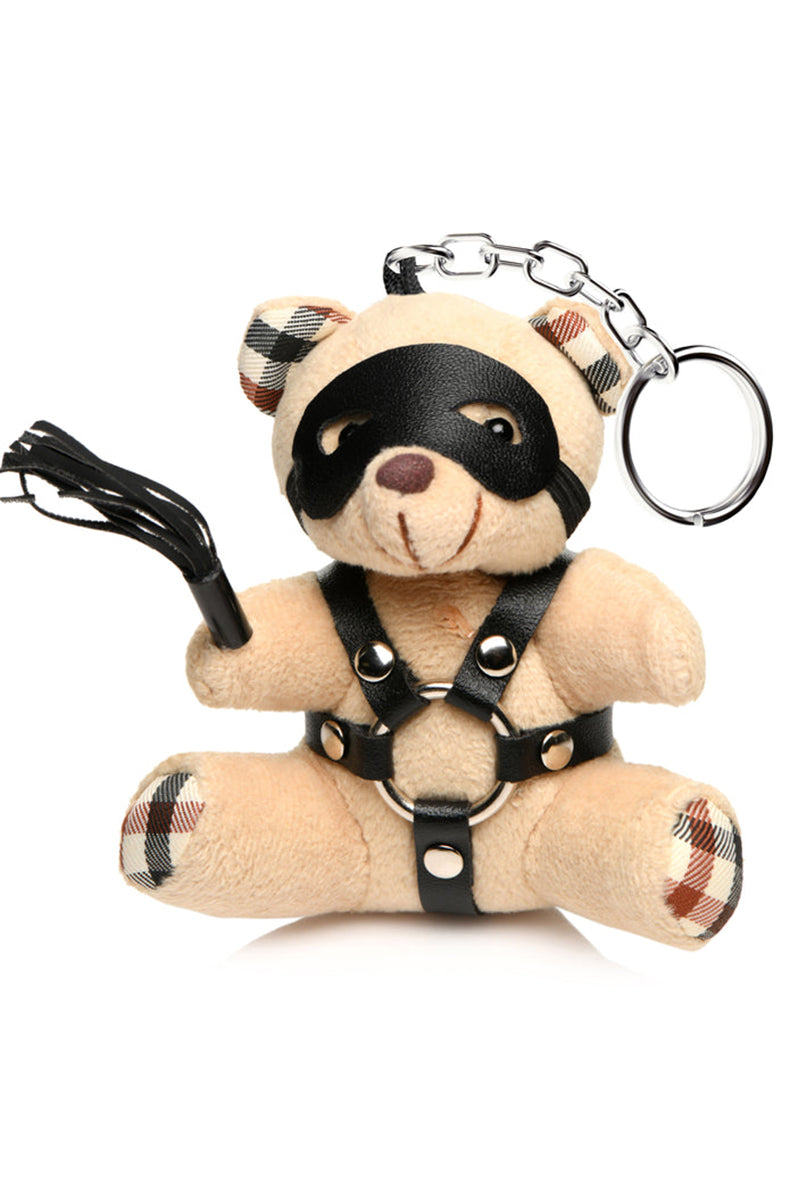 Tan plush teddy bear keychain in black PU harness with silver O-rings, black eye mask, mini flogger, and silver key ring.