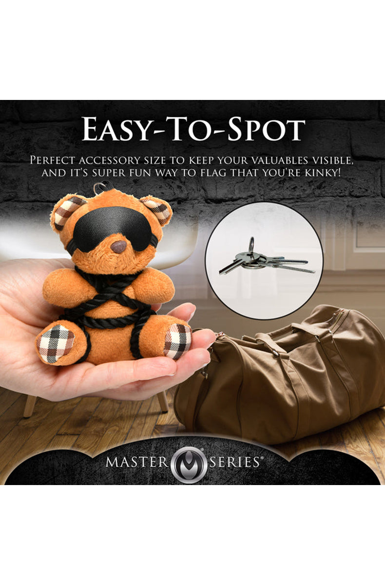 Close-up of black blindfold and shibari rope chest pattern on tan plush teddy bear keychain.