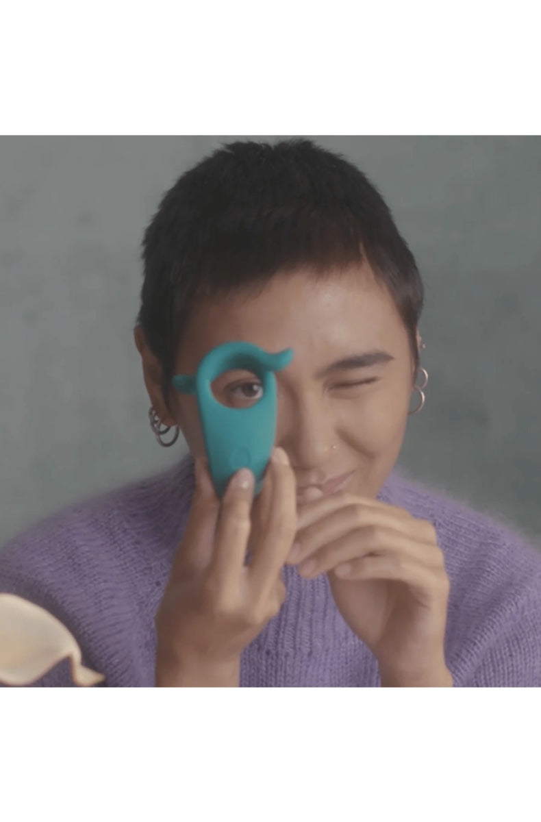 Model in purple sweater holding teal vibrating ring up to face in lifestyle setting