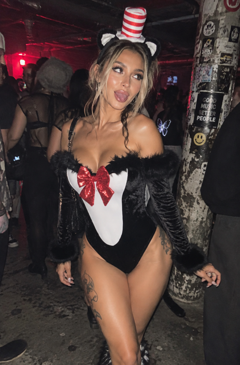 A woman posing in a black and white bunny costume with a red bow, black feathered jacket, furry boot covers, and a red and white striped hat in a dimly lit, crowded indoor setting.