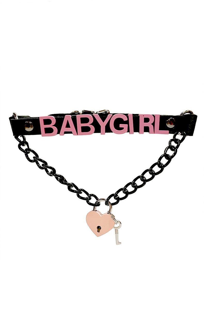 Black vegan-leather collar with "BABYGIRL" in bold pink lettering, a black chain swag draping below the front, and a pink heart-shaped padlock with a silver key hanging from the chain.