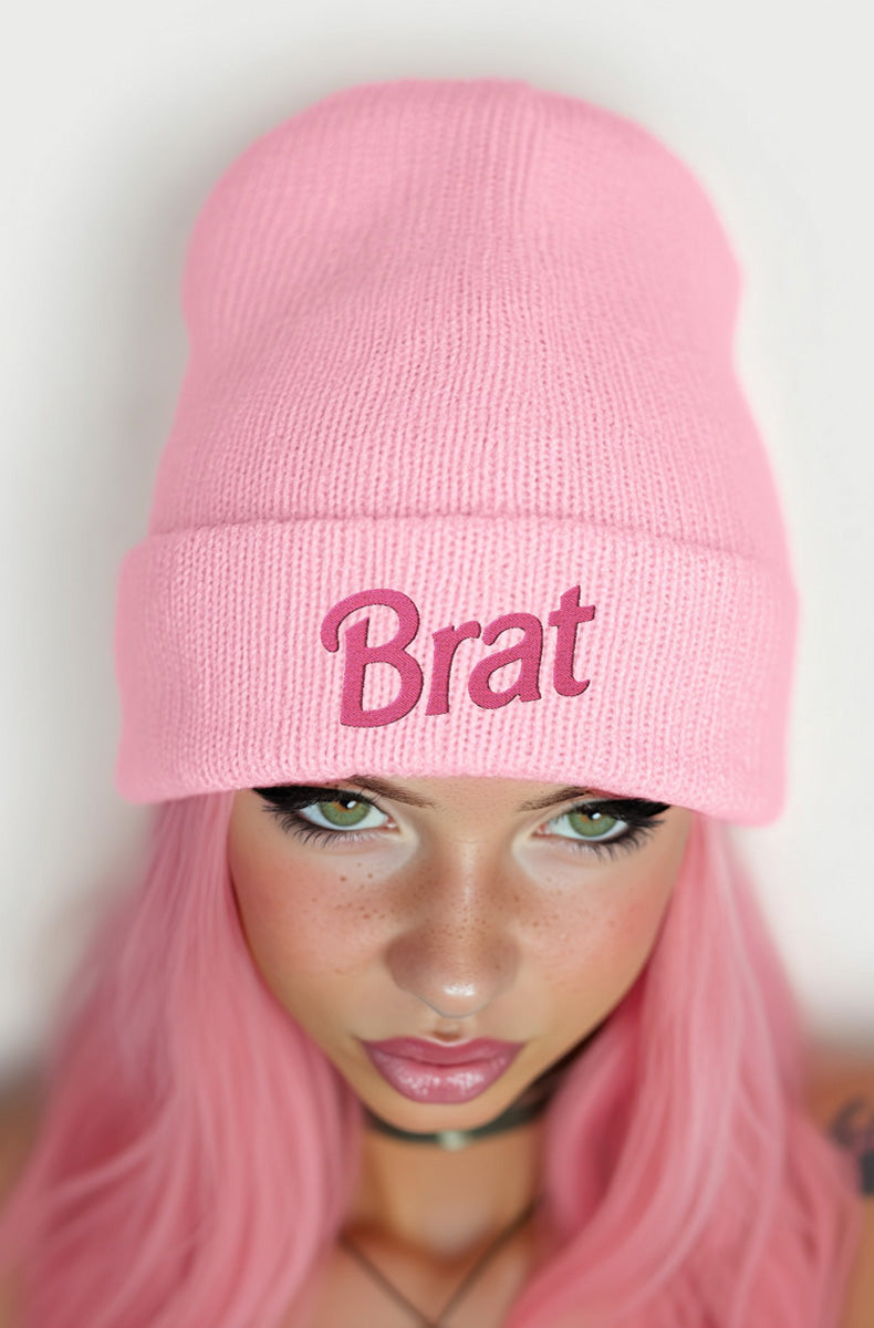 Soft pastel pink cuffed knit beanie with bold hot-pink "Brat" embroidery across the front of the cuff, worn by a model with pink hair.