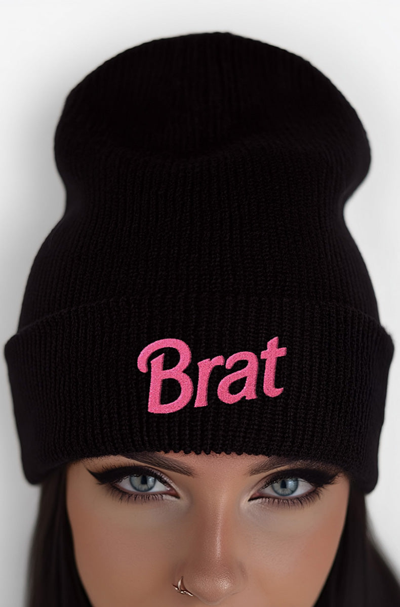 Soft pastel pink cuffed knit beanie with bold hot-pink "Brat" embroidery across the front of the cuff, worn by a model with pink hair.