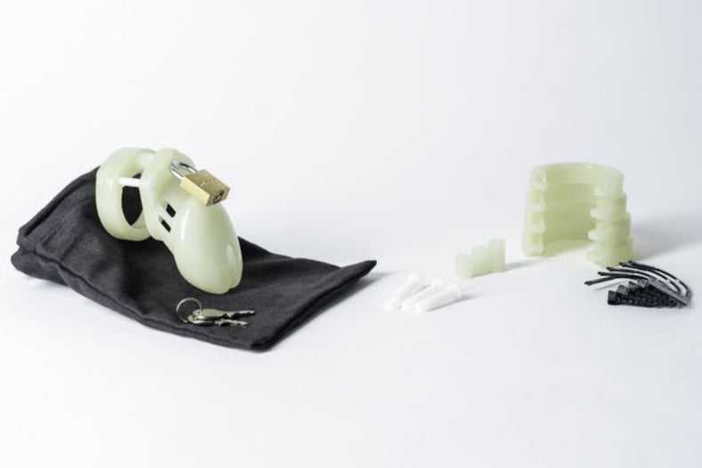 Glow chastity cage kit: pale green cage with brass padlock on a black bag, keys, five rings, pins, and numbered locks.