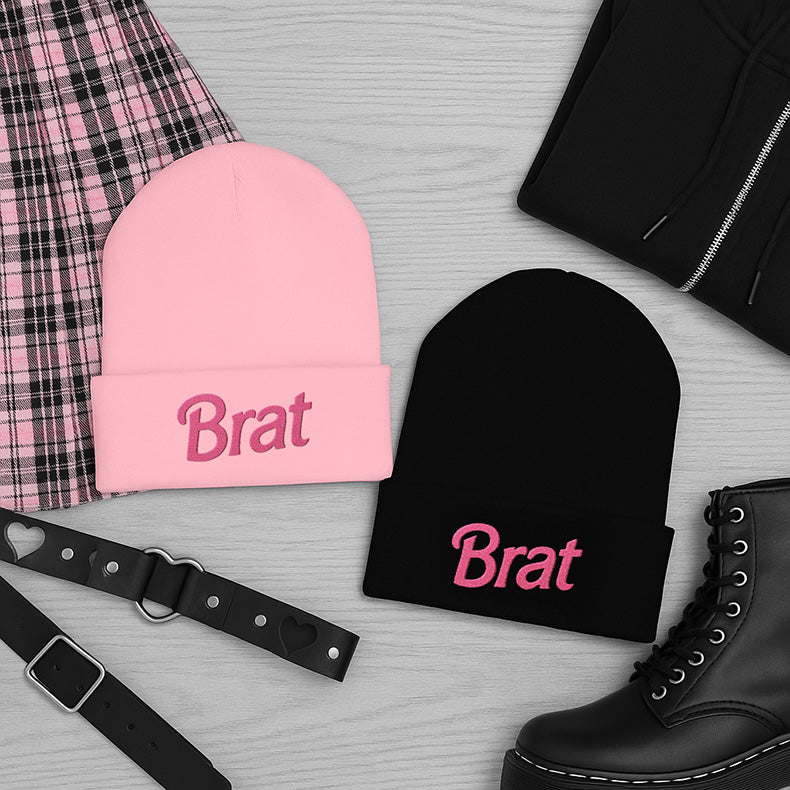 Soft pastel pink cuffed knit beanie with bold hot-pink "Brat" embroidery across the front of the cuff, worn by a model with pink hair.