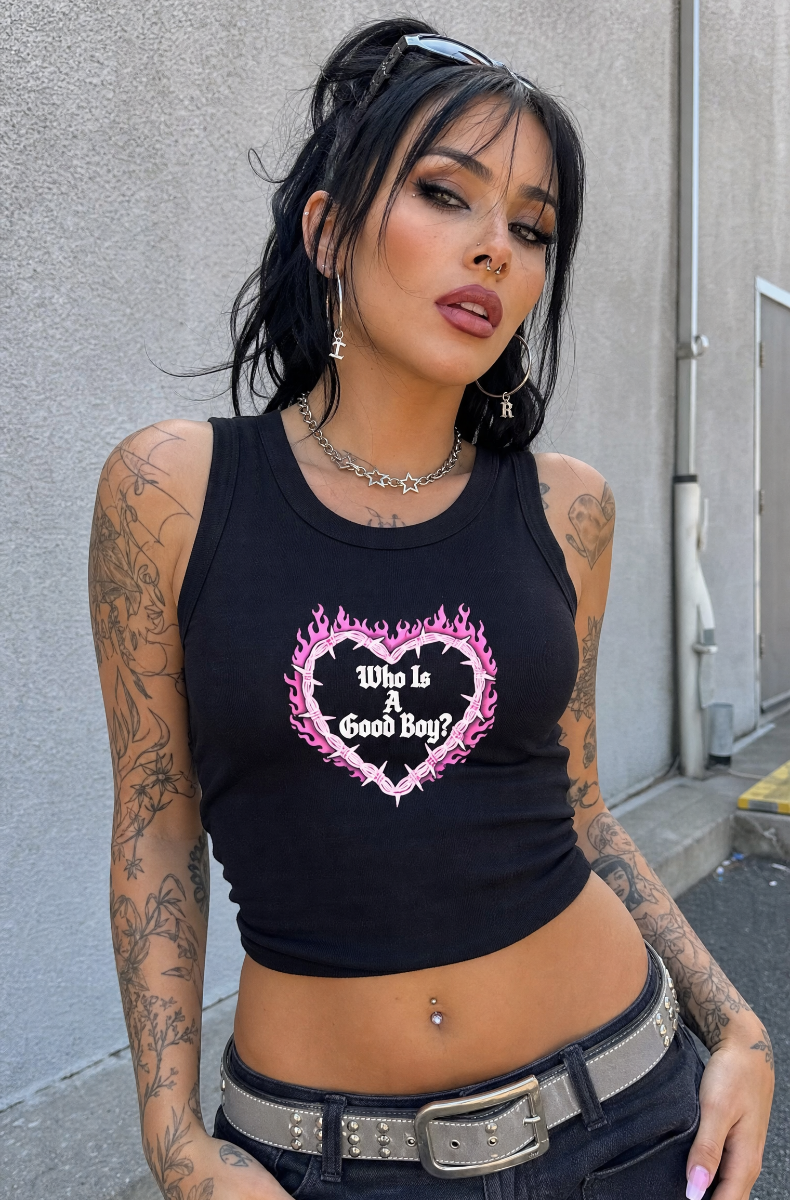 Black Who Is A Good Boy racerback tank with a pink flaming barbed-wire heart graphic, worn by a model by a wall.