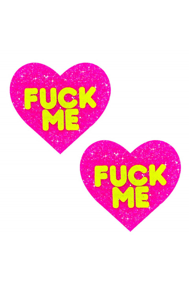 Two hot pink glitter heart-shaped pasties with "FUCK ME" printed in chunky neon-yellow block letters.