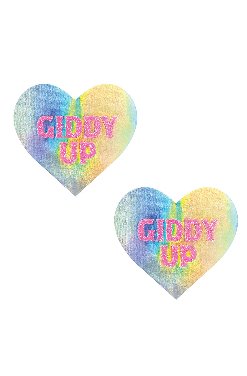Two heart-shaped pasties with a pastel rainbow tie-dye gradient and "GIDDY UP" printed in chunky pink glitter block letters.
