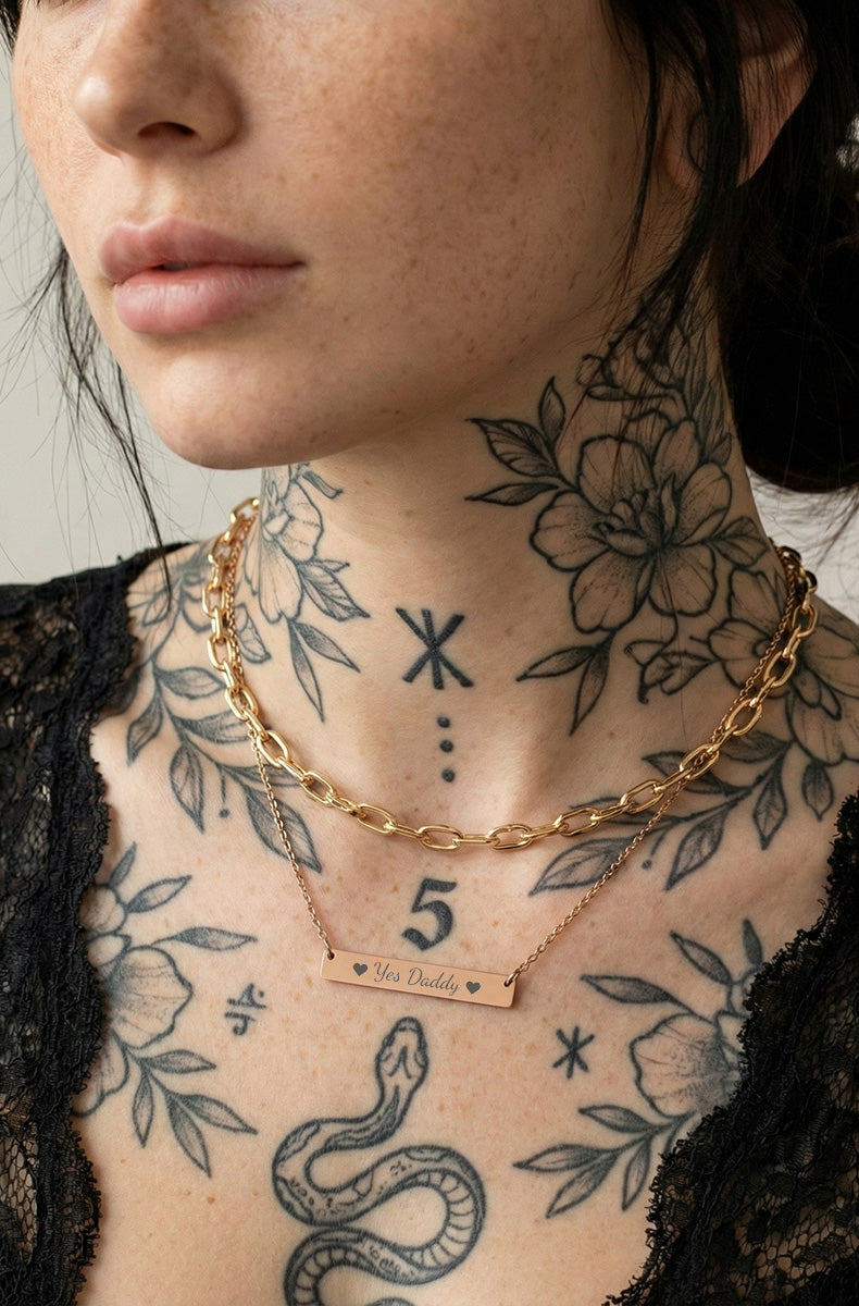 Person with tattoos wearing a gold 'yes daddy' necklace with a nameplate