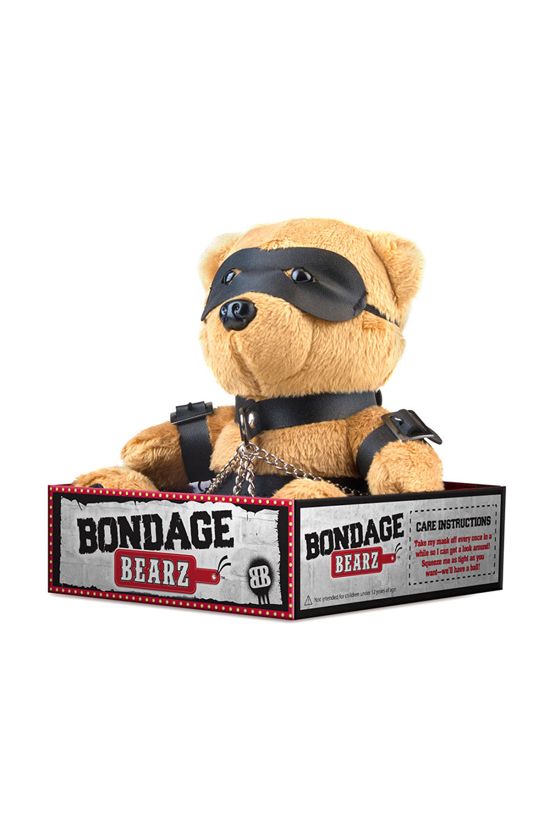 Side view of tan plush teddy bear showing black chest harness, silver chains, and arm and leg cuffs.