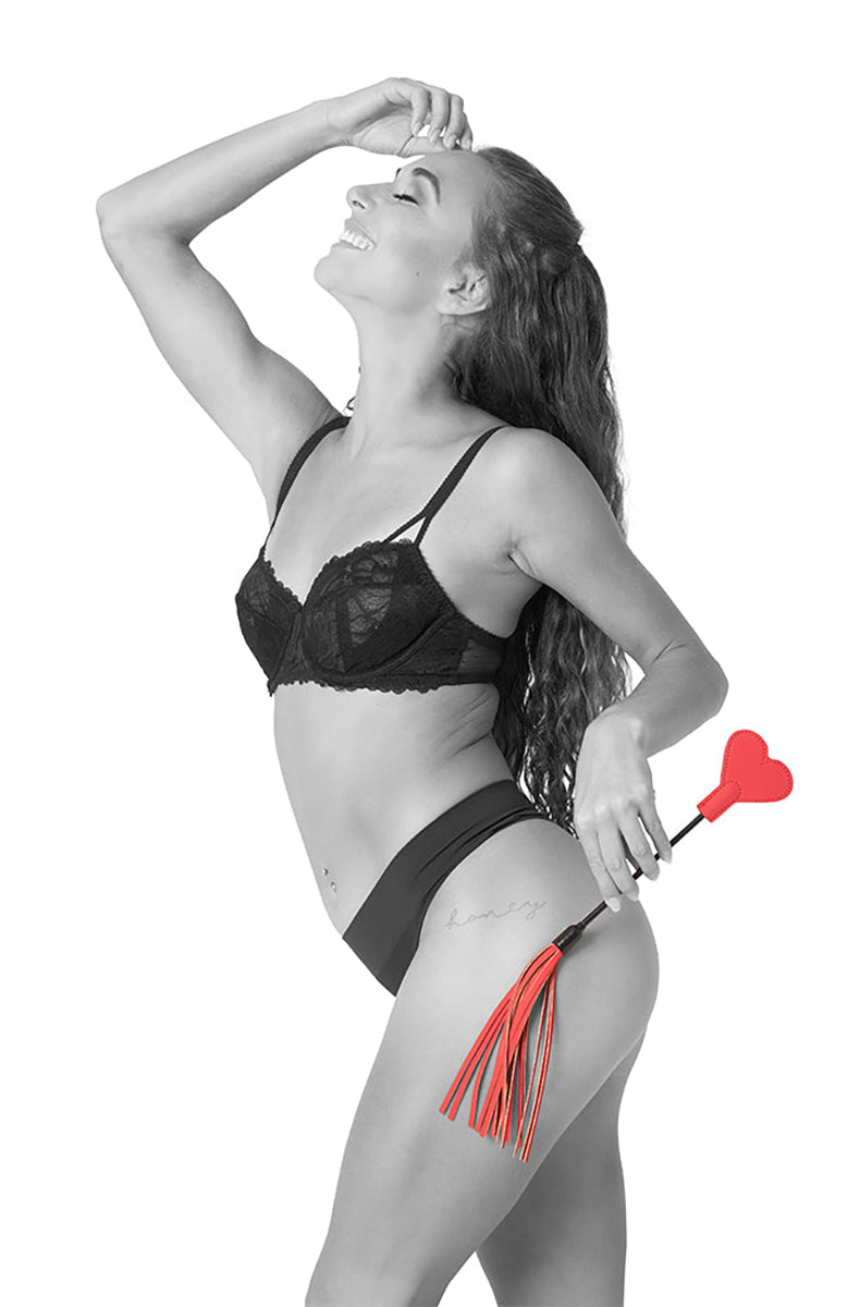 Model in black lace bra and panties holding the red heart-tipped crop with red mini-flogger tassels behind her hip.