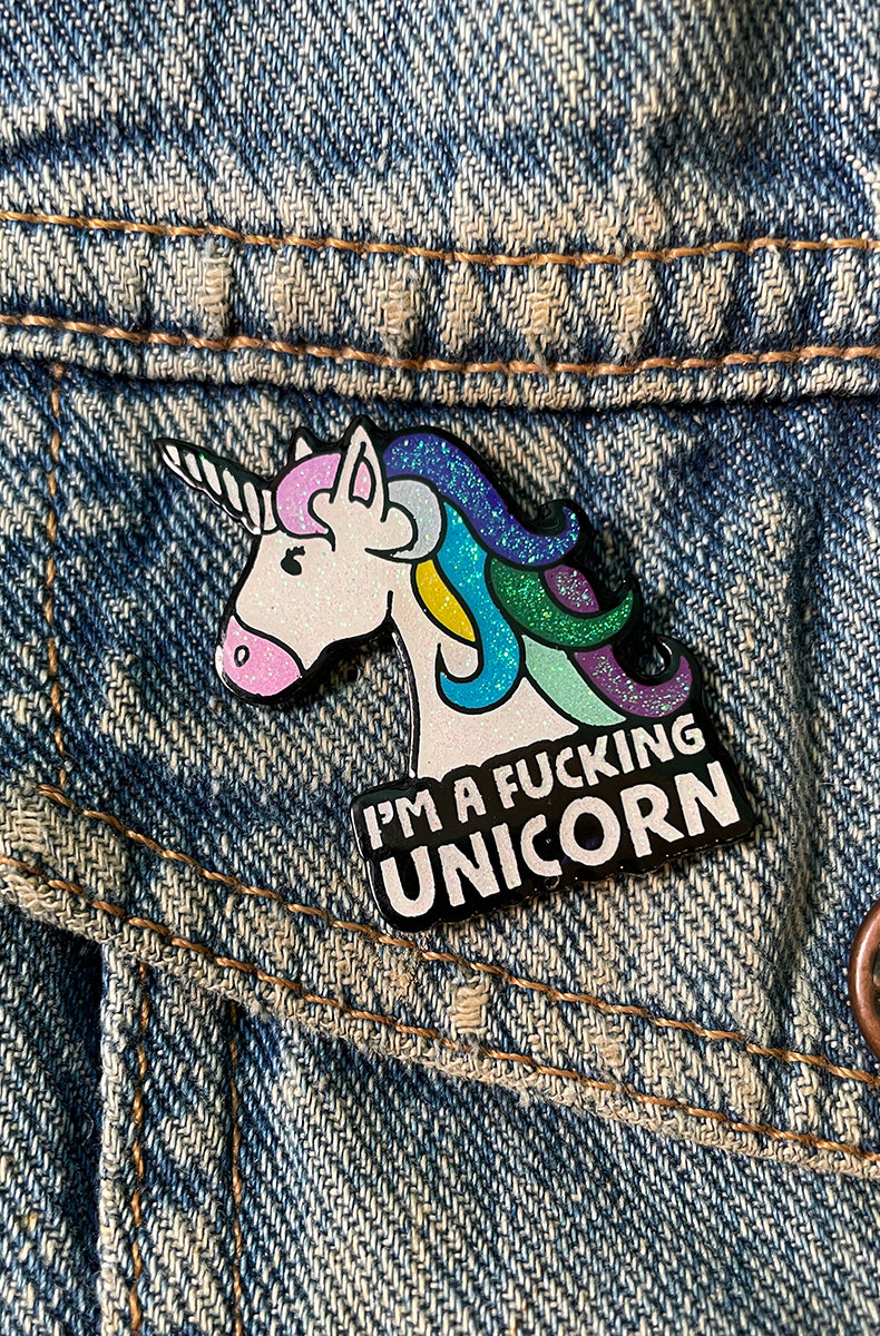 Glittery rainbow unicorn-head enamel pin paired with a black banner reading I'M A FUCKING UNICORN, attached to a denim jacket pocket.