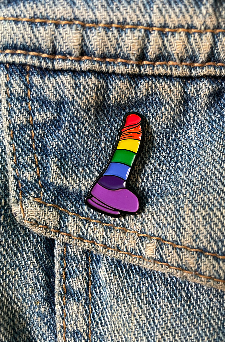 Pride-rainbow striped dildo enamel pin with red orange yellow green blue indigo bands and a purple base, attached to a denim jacket pocket.