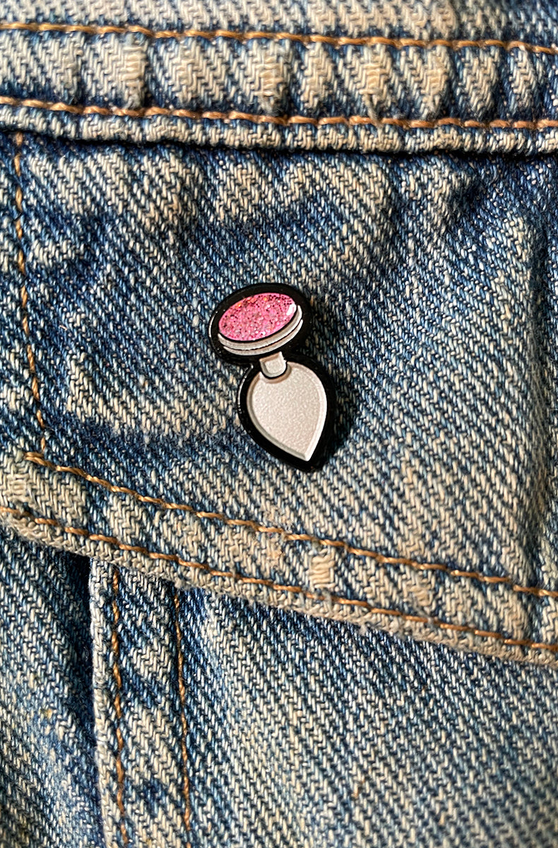 Miniature enamel pin shaped like a butt plug, white body with sparkly pink jeweled base, attached to a denim jacket pocket.