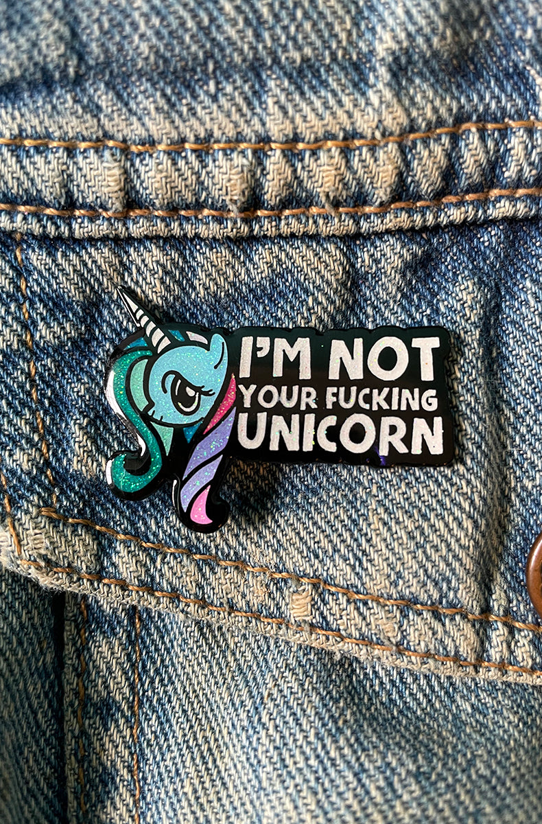 Glittery teal and purple side-profile unicorn enamel pin with a half-lidded annoyed expression next to a black banner reading I'M NOT YOUR FUCKING UNICORN.