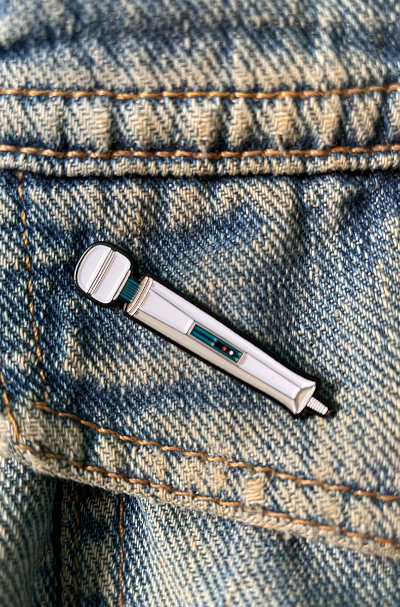 White enamel pin shaped like a classic magic-wand-style massager with a teal control window mid-handle, attached to a denim jacket pocket.