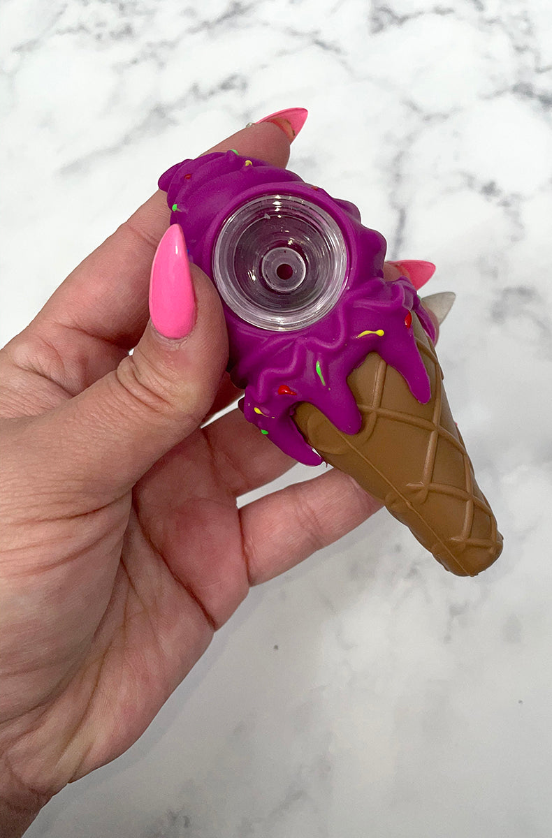 Three PIECEMAKER ice cream cone silicone pipes in yellow, purple, and pink with sprinkle accents and glass bowls, on marble.