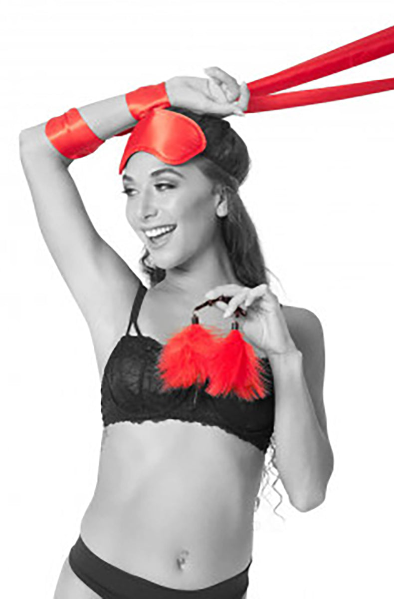 Model wearing the red satin blindfold with arms raised, tethers wrapped around wrists, holding feather clamps.