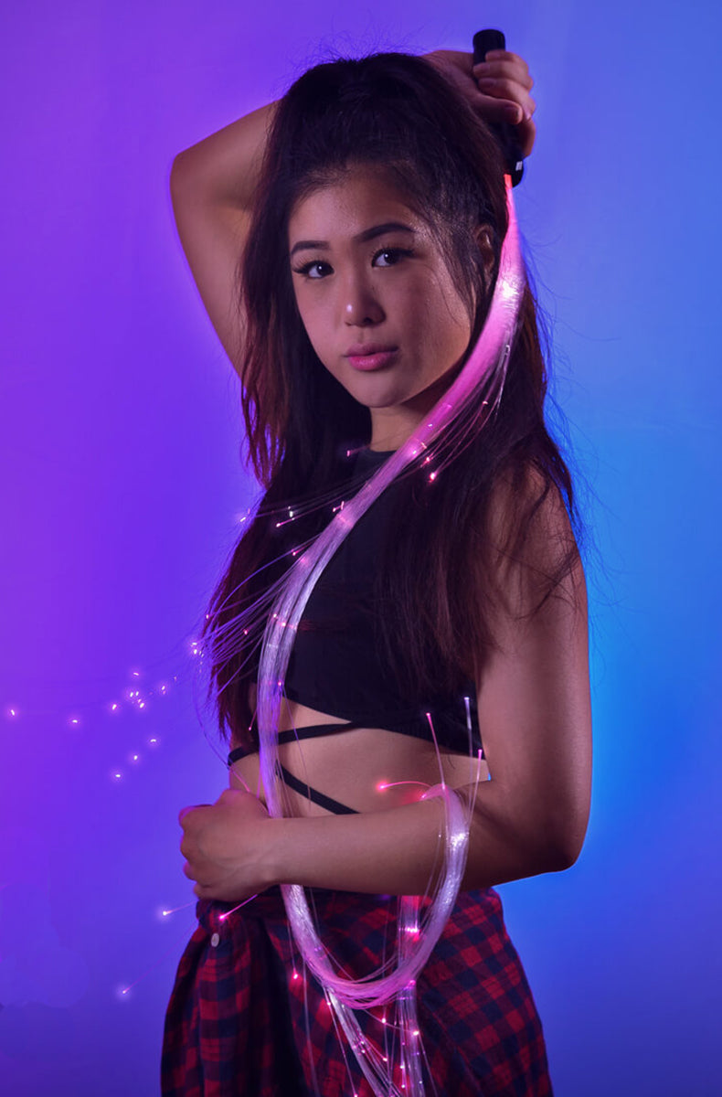 Person wearing black holding a long fiber-optic LED whip with pink and white glowing fibers trailing in the air against a purple-blue lit background.