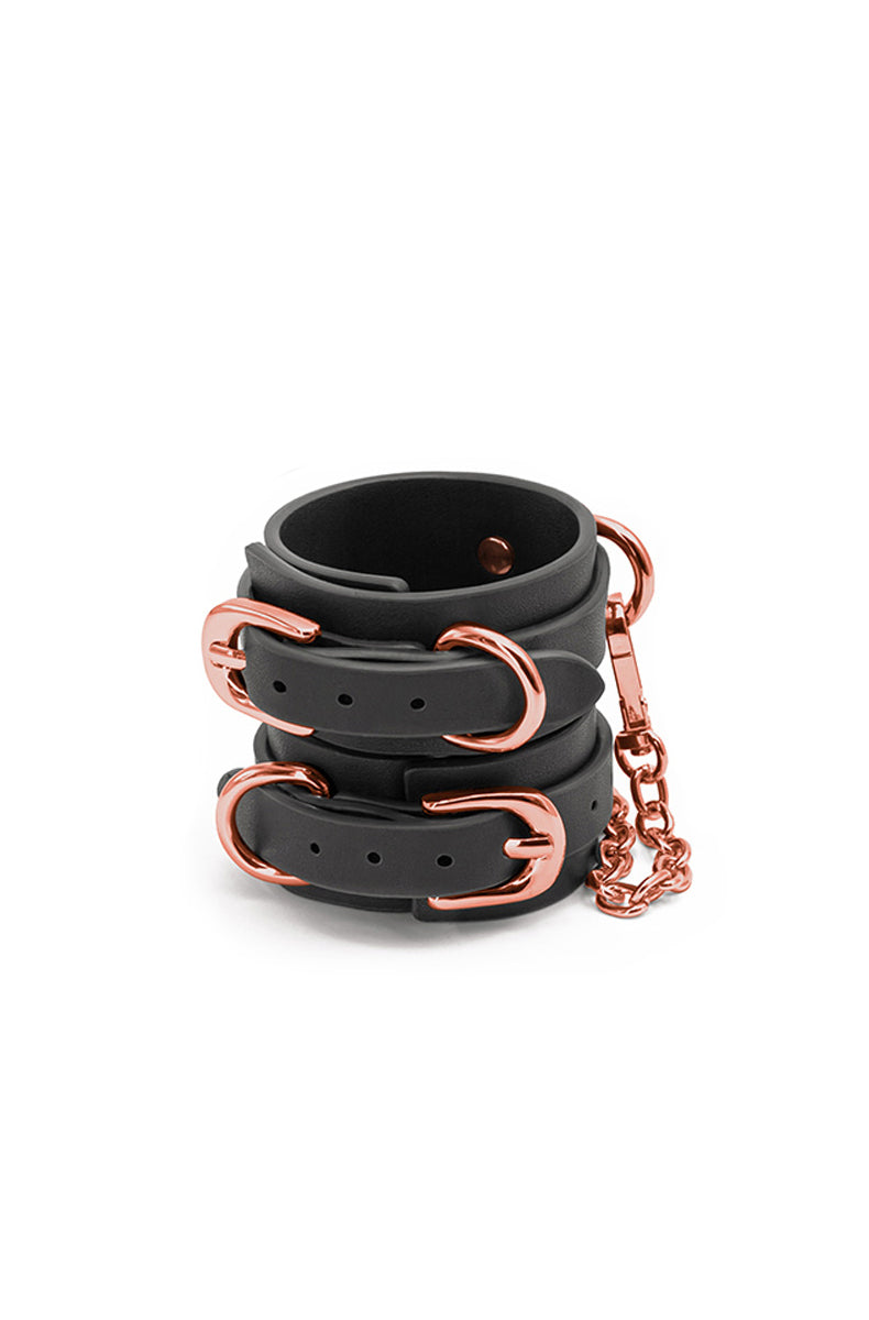 Stacked pair of black synthetic-leather wrist cuffs with rose-gold buckles, D-rings, and a rose-gold connecting chain.