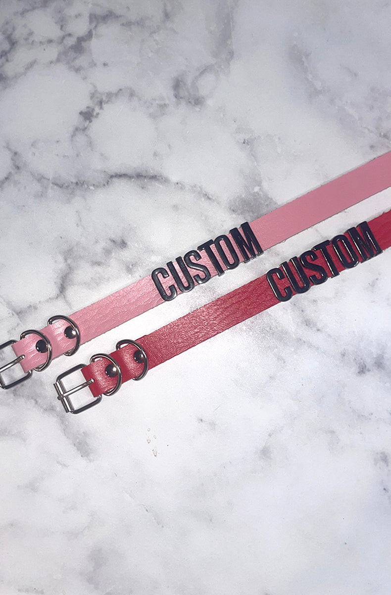 Pink and red vegan leather collars with raised black plastic letters spelling CUSTOM as a sample, both with silver buckles and D-rings.