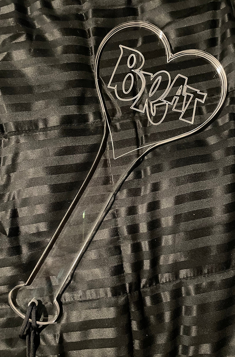Front view of the clear acrylic BRAT heart paddle on a black striped satin background with a black wrist cord.