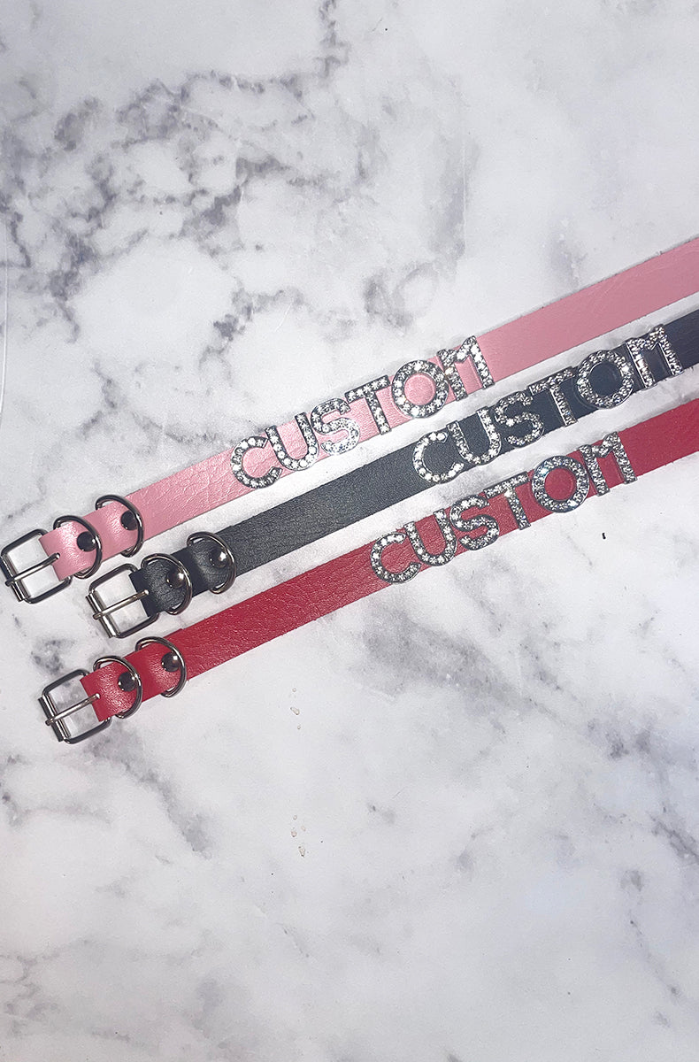 Pink, black, and red vegan leather collars with silver rhinestone-encrusted letters spelling CUSTOM as a sample, all with silver buckles.