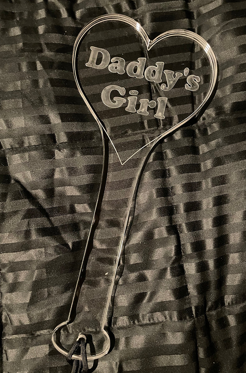 Front view of the clear acrylic Daddy's Girl heart paddle on a black striped satin background with black wrist cord.