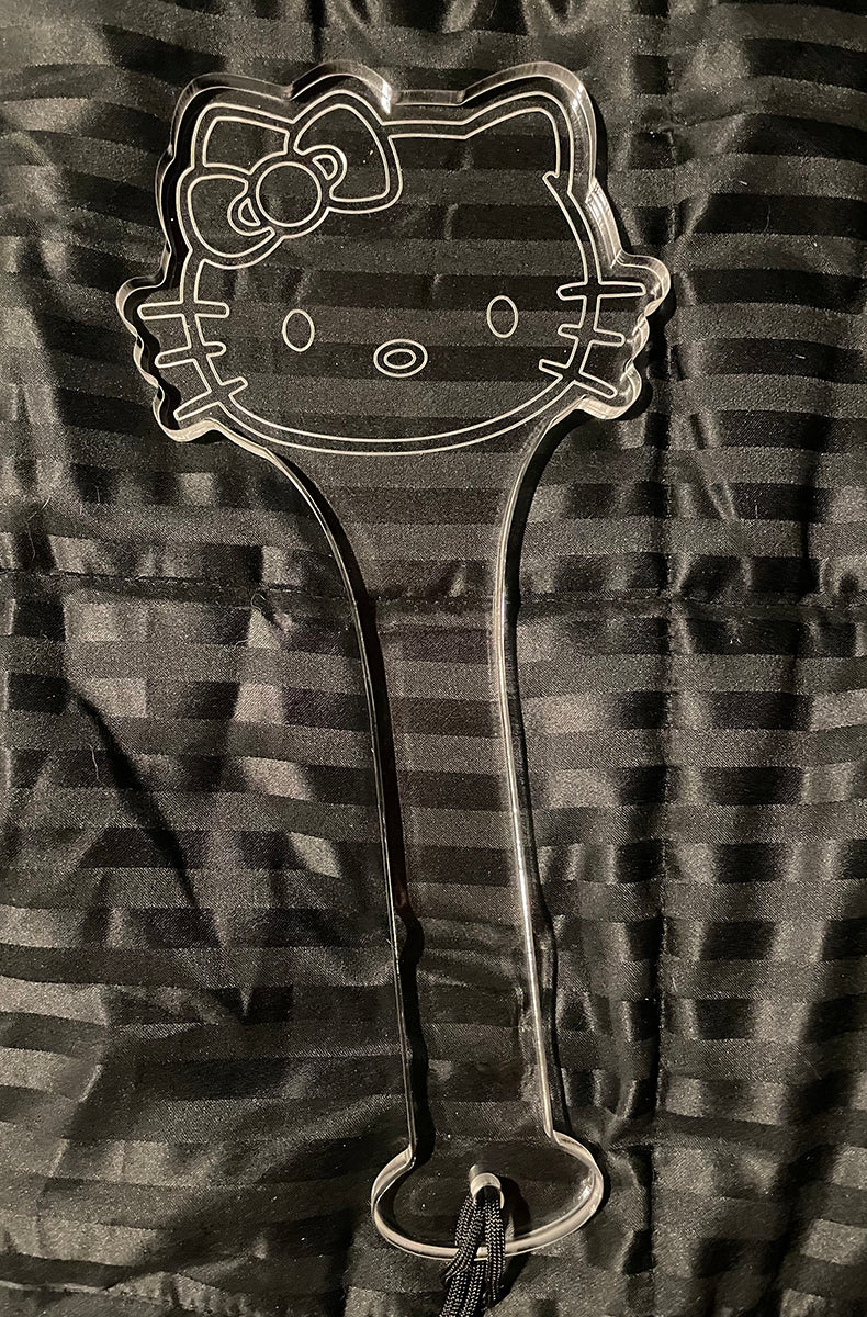 Front view of the clear acrylic kawaii cat paddle laid flat on a black striped satin background with black wrist cord.