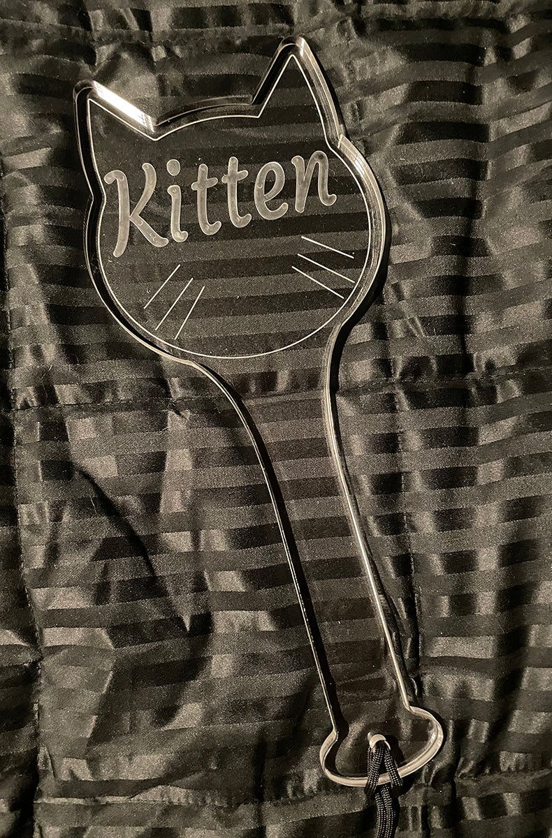 Front view of the clear acrylic Kitten paddle laid on a black striped satin background with a black wrist cord and ring.