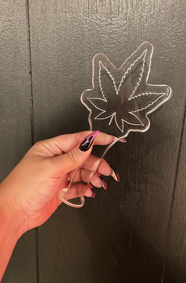 Clear acrylic cannabis-leaf paddle held in a hand with pink and black nails against a dark wood wall.