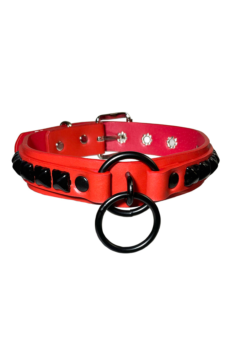 Red vegan-leather collar with rows of glossy black pyramid studs along both sides and twin black O-rings at the front center.